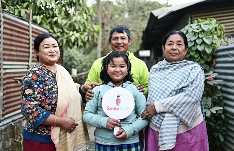 Bena smiling and holding a Smile Train sign with her parents and grandmother after cleft surgery