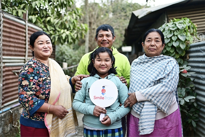Bena smiling and holding a Smile Train sign with her parents and grandmother after cleft surgery